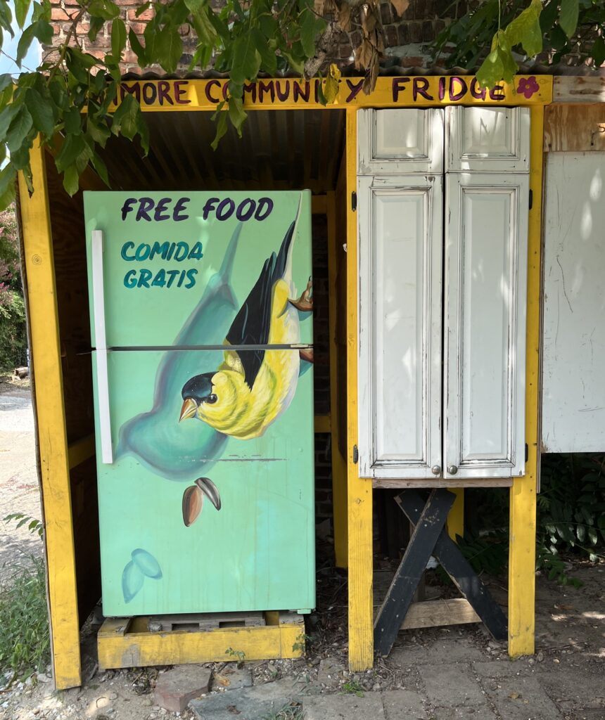 Standing outside beneath the low hanging branches of a leafy tree, a mint-colored fridge featuring a painting of a yellow and black bird says: “Free food.”