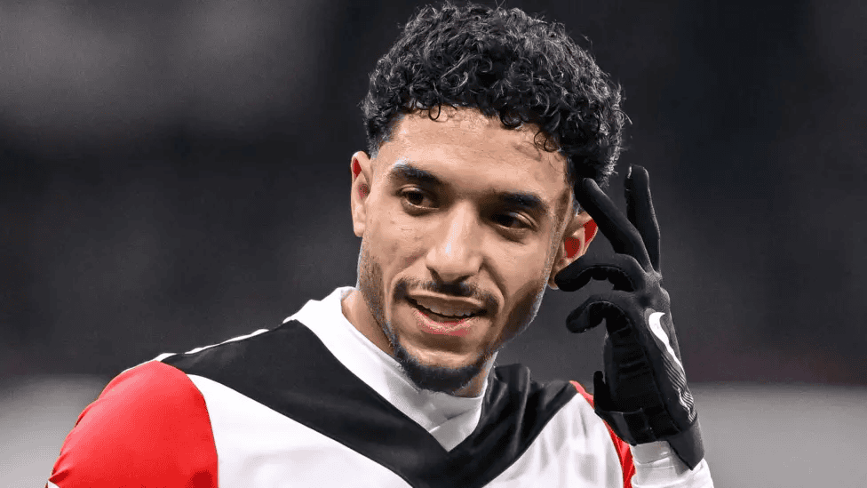 A man with curly hair wearing a Real Madrid jersey and gloves adjusts his earpiece on the sports field, his expression focused, as if receiving a crucial weekly update.
