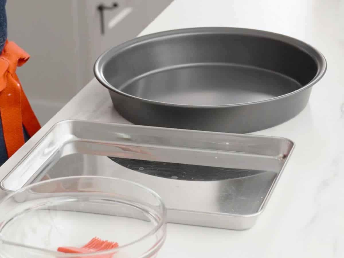 A round nonstick cake pan, a rectangular stainless steel tray, and a bowl with oil and a pastry brush in it.