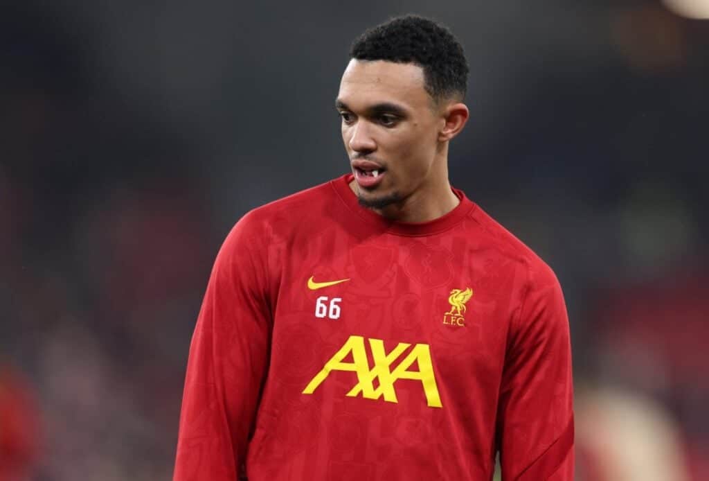 A Liverpool defender in a red football jacket stands on the field, glancing to the side.
