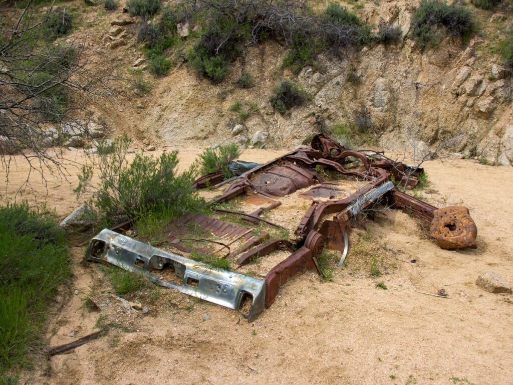 Landscape image of the rusted remains of an old car in the middle of the wash on the Blue Wash Hiking Trail, near Carefree, Arizona. Grasses, shurbs and hillsides in the background, Easy Hiking Trails, Central Arizona, Northern Phoenix Area, Family and Dog Friendly Hiking Trails, Copyright azutopia.com.