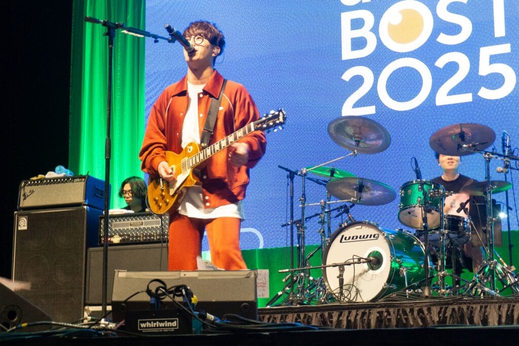 Photograph of Shunichi Tanabe from Blue Encount. He's wearing a white shirt and and orange jacket and pants. He's singing into a microphone on stage while playing guitar in front of a sign that reads "ANIME BOSTON 2025"
