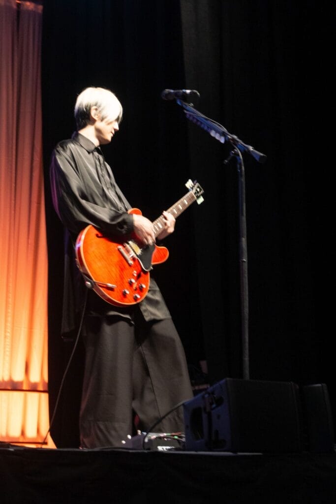 Photograph of Blue Encount guitarist Yuya Eguchi playing guitar onstage. He's wearing an all-black ensemble as he leans back