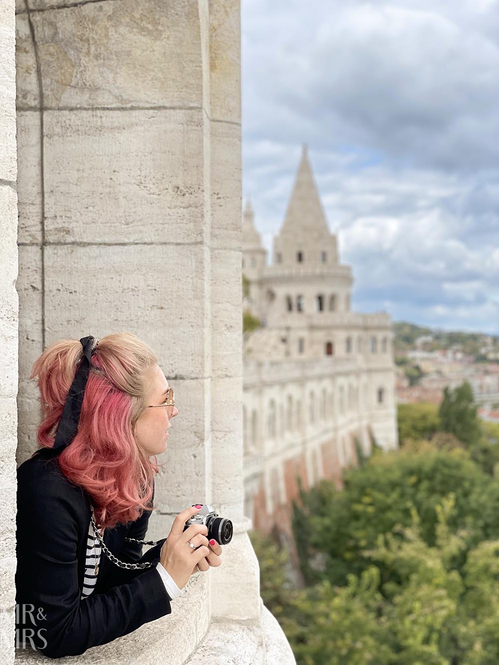 Essential travel guide to Budapest, Hungary - Christina looking out of the castle walls of Fisherman's Bastion. Woman with pink hair and camera leaning out over ancient walls of castle