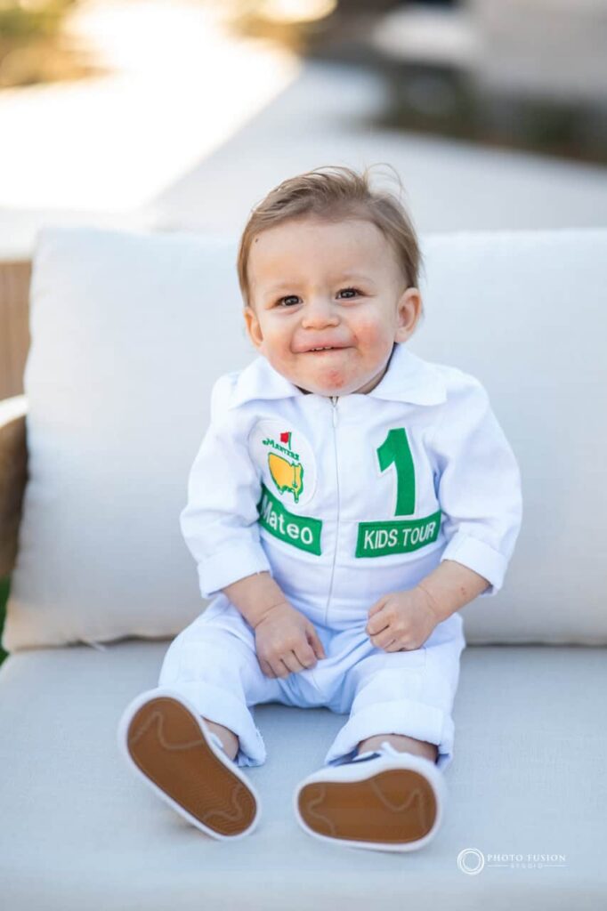 Event Photography for an Unforgettable First Birthday 25 This is an image of the child who is turning one years old at his birthday party. He is wearing a custom caddie outfit that has the masters logo on the from and his name next to it. The is a giant one on the front as well that says kids tour under it.