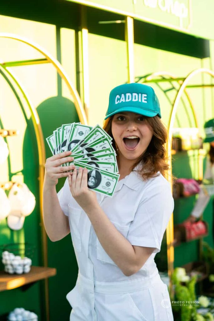 Event Photography for an Unforgettable First Birthday 39 This is a lady who is at a one year olds birthday party, she is wearing a white caddie outfit and a green hat that has the word caddie on it. She is looking at the camera while posing and holding fake money with the childs face who is turning one.