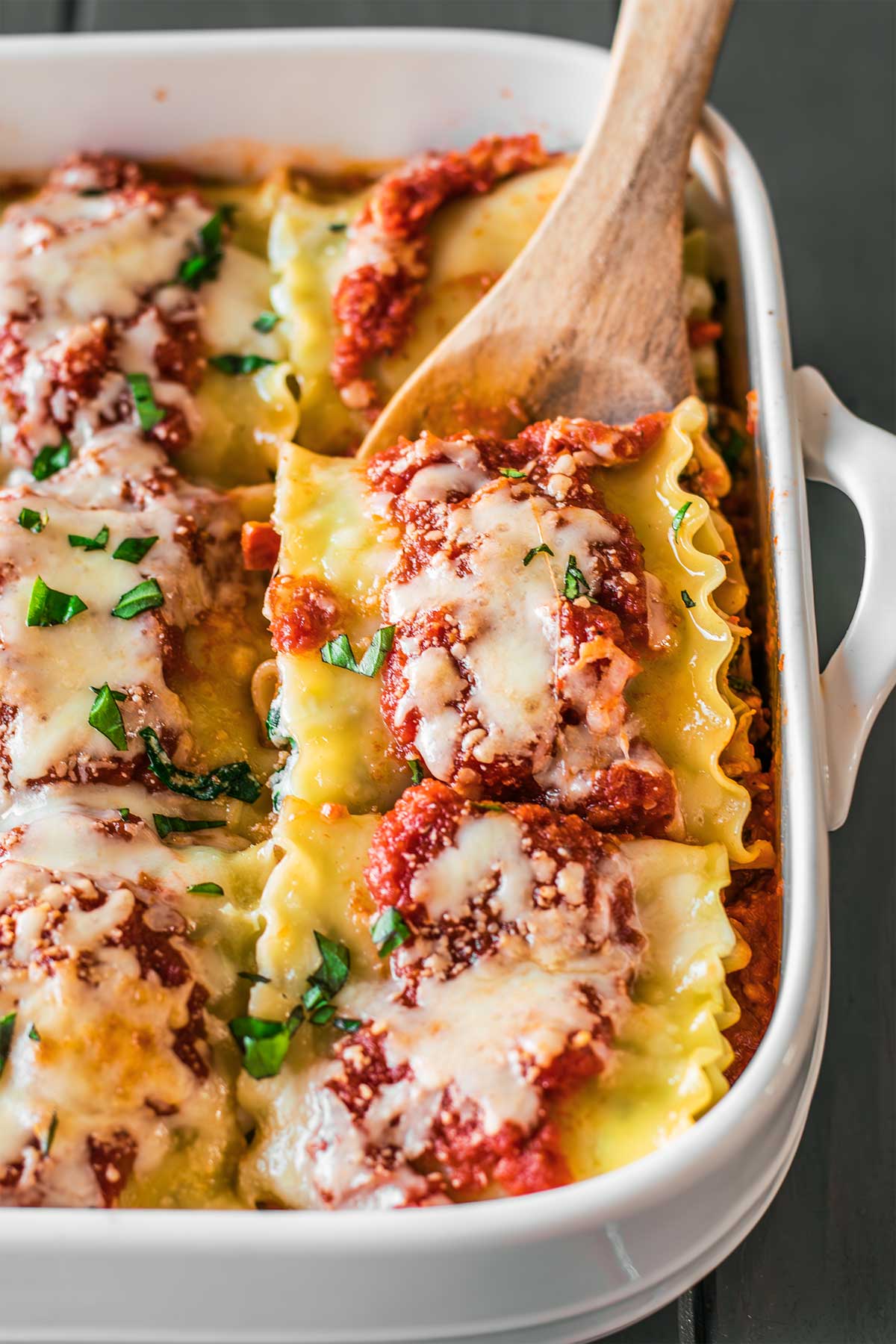 Close up top down view of a casserole dish filled with rows of freshly made lasagna roll ups, pictured with a wooden spoon dipping behind a roll up, prepared to lift it out of the dish.