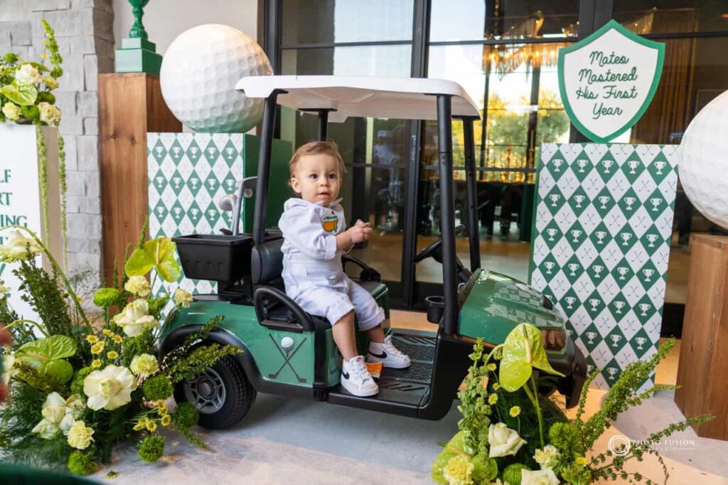 Event Photography for an Unforgettable First Birthday 1 A child is sitting in his mini golf cart that is made for kids and smiling. He is celebrating his first birthday that is a masters golf theme party. There are flowers on both sides of the cart and giant golf ball displays as well.