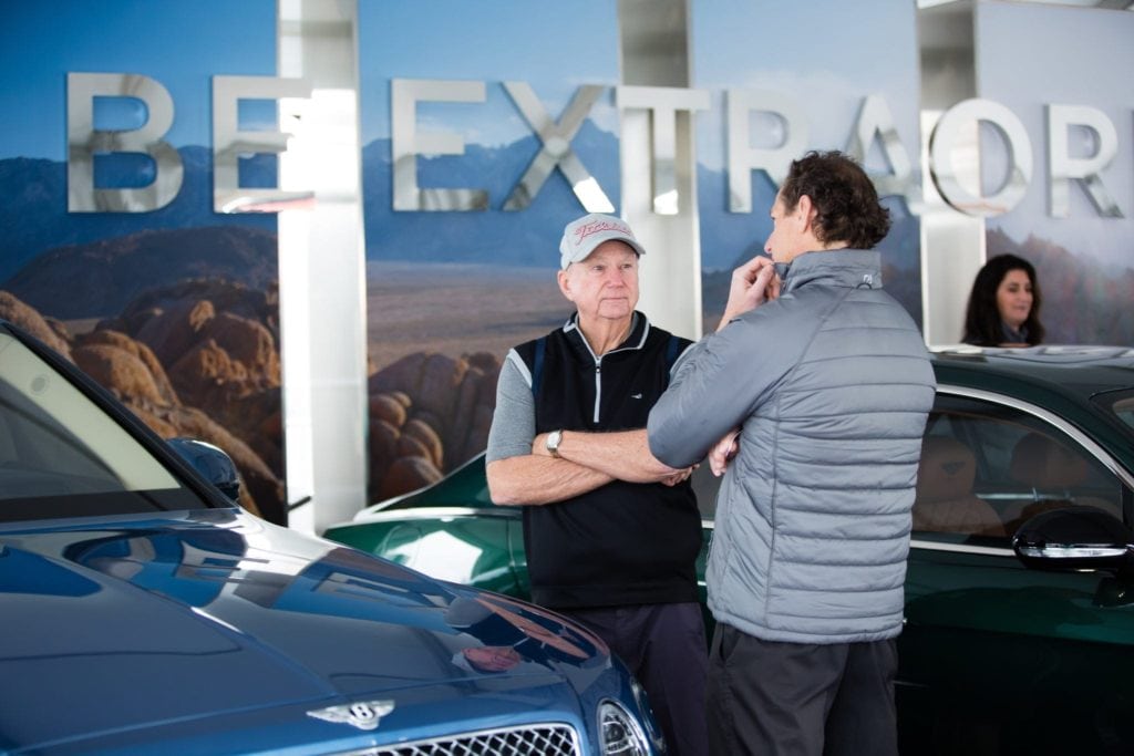 Two men stand between luxury cars, one in a jacket and the other in a hat, conversing at a corporate event. A large sign with "BE EXTRAORDINARY" is partially visible in the background, surrounded by other attendees and a captivating landscape image—a snapshot of exquisite event photography.