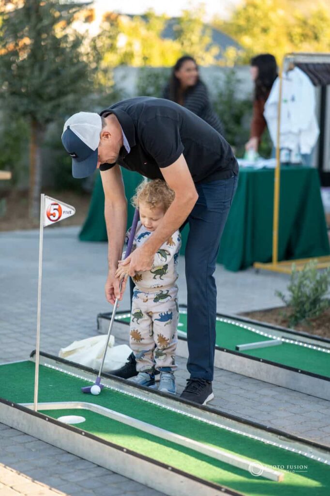 Event Photography for an Unforgettable First Birthday 8 Dad and son on a mini putting corse at a one yearolds birthday party. The dad is teaching his sone how to hold a golf club and play mini golf.