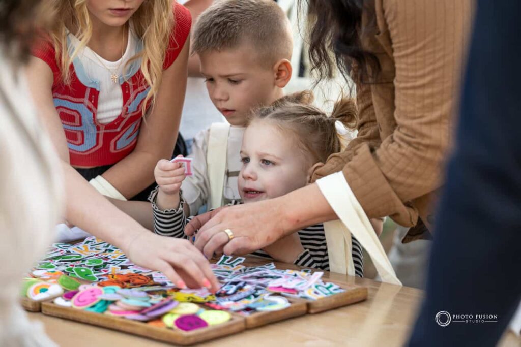 Event Photography for an Unforgettable First Birthday 29 A group of people are gathered around a table and picking patches to make their custom tshirts with. There is a little girl in the center who is holding a patch and looking at it with a smile on her face. Hands can be seen around her reaching for them.