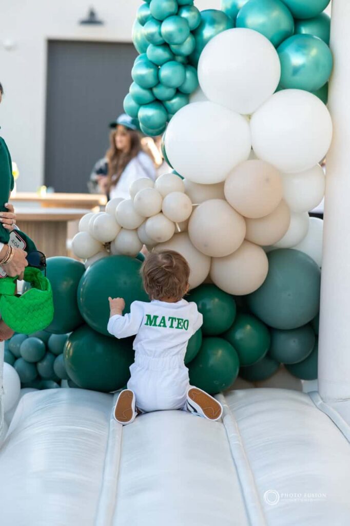Event Photography for an Unforgettable First Birthday 26 This is an image of the birthday boy at his bounce house with his back to the camera. His caddie outfit reads his name and he is playing with the green balloons that are at the bounce house.