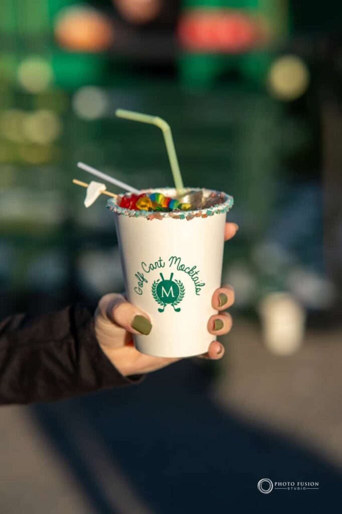 Event Photography for an Unforgettable First Birthday 14 This is an image of a mocktail that a lday is holding. The mocktail has a rock suger rim, a toothpick with gummy bears sticking out of it, and a green straw. The cup has a custom design on it that is green and has an M in the middle with golf clubs on the sides.