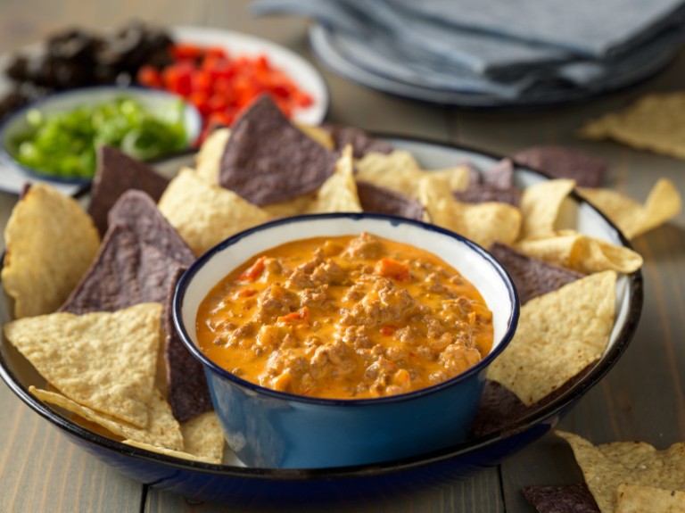 Cheese dip with salsa and beef in a blue bowl on a plate with blue and white corn tortilla chips. 