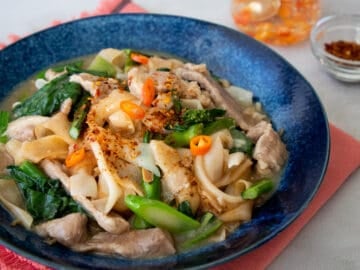 a blue bowl of rad na with chili vinegar and chili flakes on the side