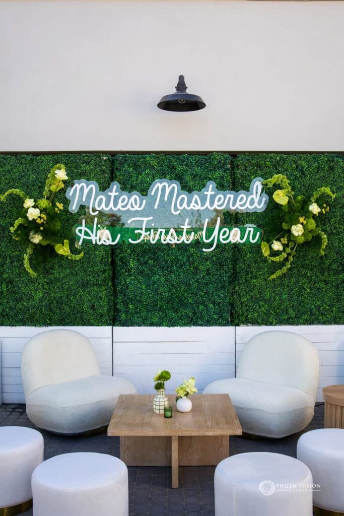 Event Photography for an Unforgettable First Birthday 15 Neon sign that says mateo mastered his first year, with a green bush panel behind it and florals comming out of the panel on both sides of the sign. There are white chairs and tomans that surround a brown table with floral decor on it.