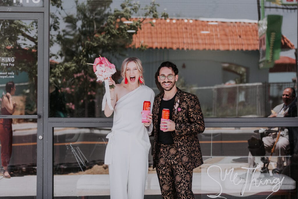 A bride and groom stand together, excitedly celebrating their wedding day, with pink beverages and a pink bouquet outside of Sure Thing Chapel in Las Vegas.