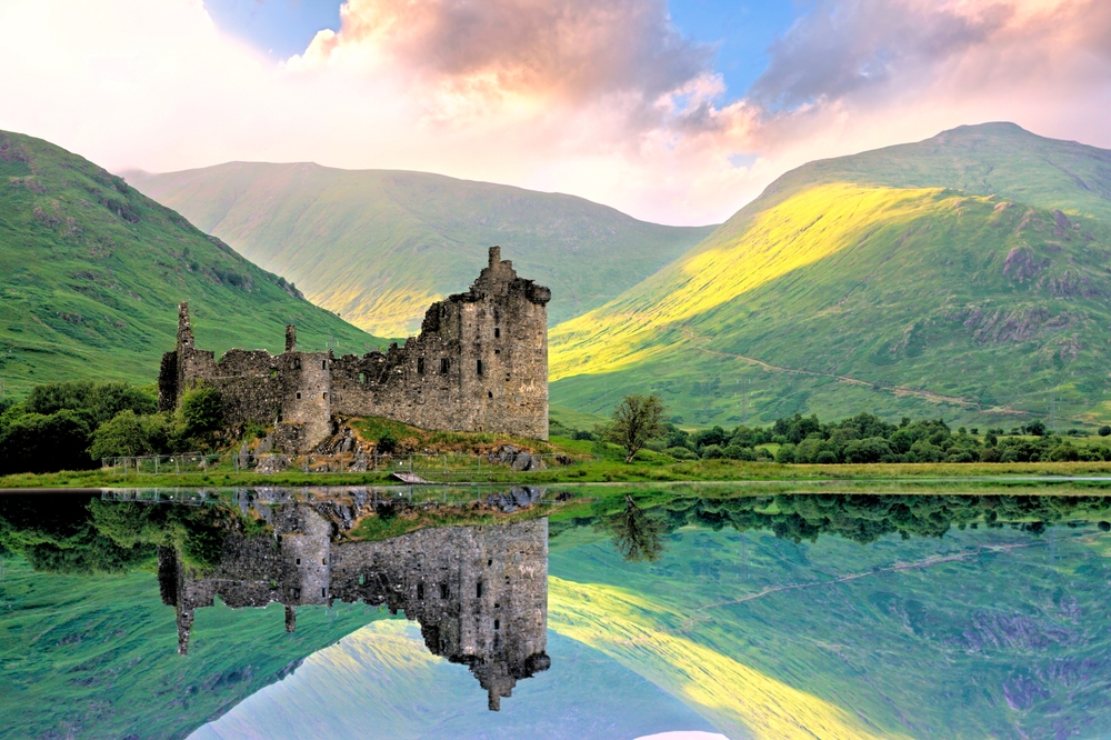 In Scotland, an hold historic and worn down castle made of stone stands tall yet weathered, it's reflection showing in the lake below, green rolling hills in the background.