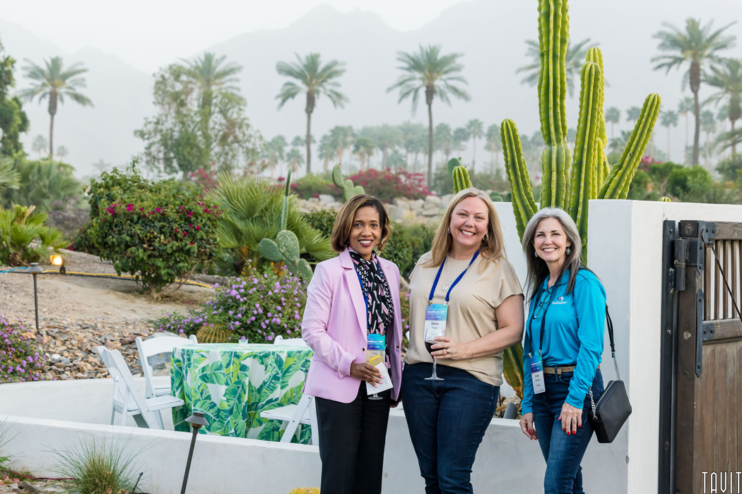 three ladies smiling outside at a corporate event in Palm Springs, CA. Beautiful backdrop of a dining patio, shrubs, cactus and palm trees