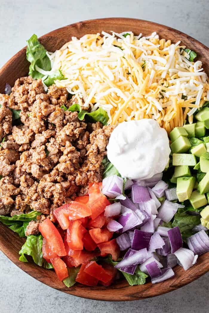 overhead view of turkey taco salad in a wooden bowl with sections of ground turkey, cheese, chopped avocado, chopped tomato, chopped onions and sour dream in the middle, all on a bed of lettuce