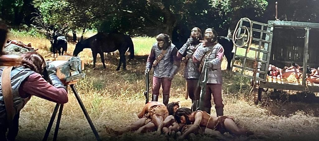 Still from Planet of the Apes depicting three gorillas holding guns and posing for the camera. Dead humans lay at the feet and in cages behind them. 